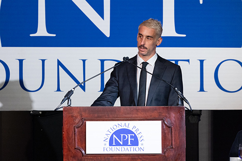 Ilie speaking at a National Press Foundation podium in front of a large event backdrop.