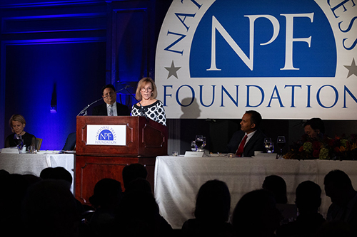 Patricia speaking at a National Press Foundation podium with others seated onstage.